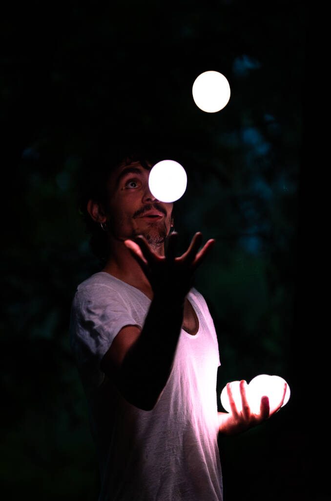 Malabarista en la oscuridad de la noche lanzando al aire tres esferas que emiten luz blanca.