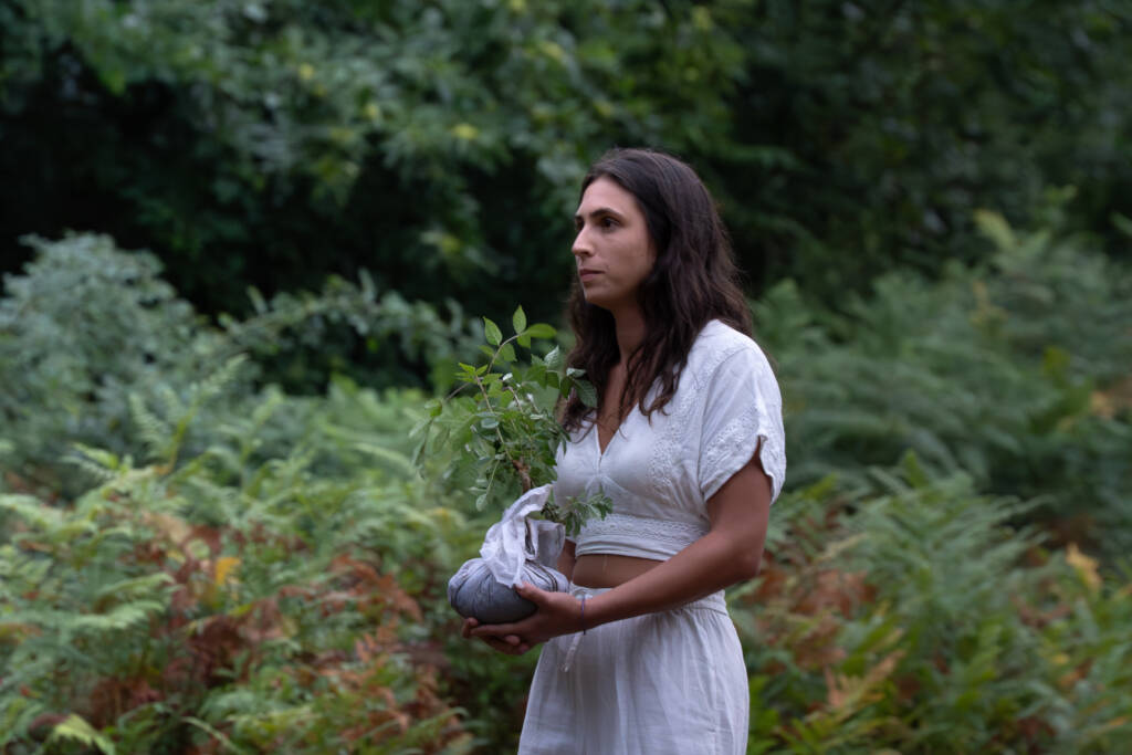 Mujer joven con el pelo oscuro y vestida de blanco sosteniendo con cuidado un pequeño árbol con las raíces envueltas en tela, rodeada de helechos y vegetación verde.