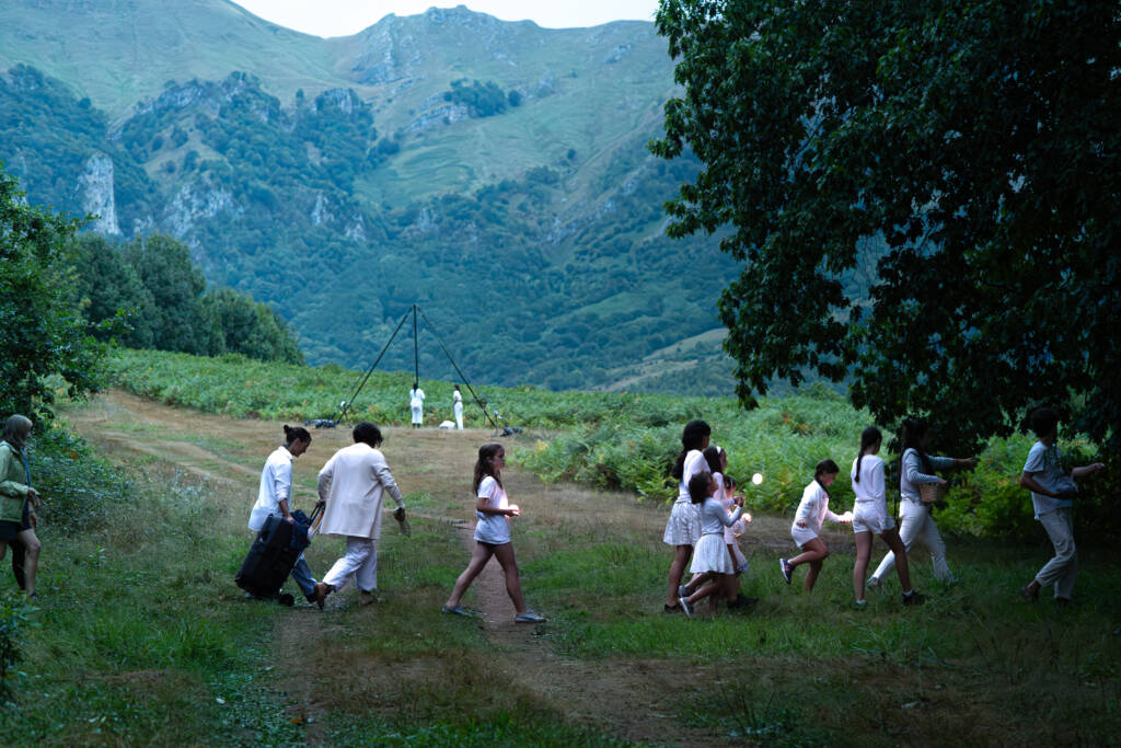 Fila de artistas y niños vestidos de blanco caminando por un prado verde llevando esferas de luz hacia una estructura piramidal, con montañas de fondo al atardecer.