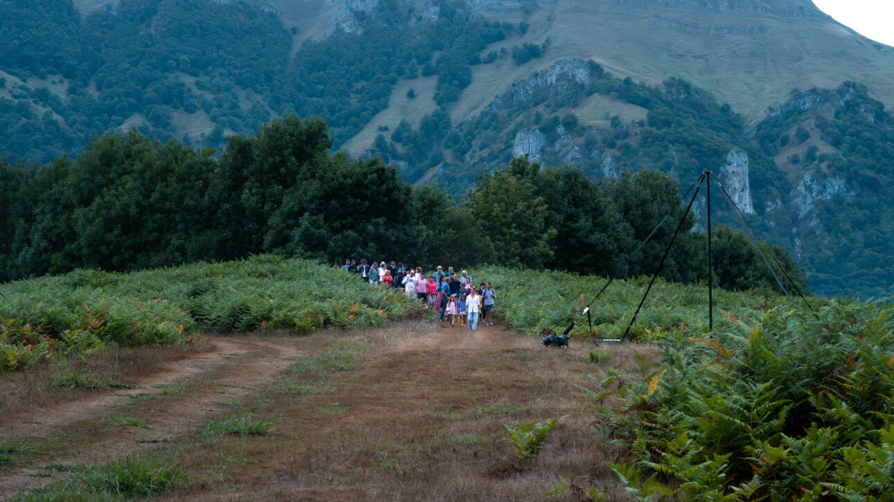 Fotografía de paisaje del Valle de Araiz. Plano general en el que se ve un grupo de 50 personas descendiendo por un camino de montaña hacia el claro que tienen delante. A los lados de las personas y del camino, hay una manta extensa de helechos. Detrás de las personas, en el el fondo de la imagen, hay montañas muy altas que tapan casi por completo el fondo de la imagen. En la parte superior, el cielo se ve blanco y encapotado por las nubes.
