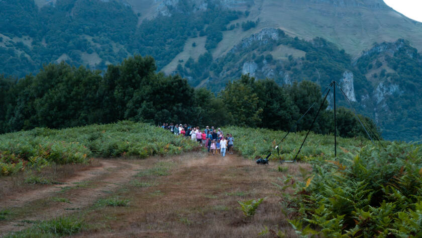 Fotografía de paisaje del Valle de Araiz. Plano general en el que se ve un grupo de 50 personas descendiendo por un camino de montaña hacia el claro que tienen delante. A los lados de las personas y del camino, hay una manta extensa de helechos. Detrás de las personas, en el el fondo de la imagen, hay montañas muy altas que tapan casi por completo el fondo de la imagen. En la parte superior, el cielo se ve blanco y encapotado por las nubes.